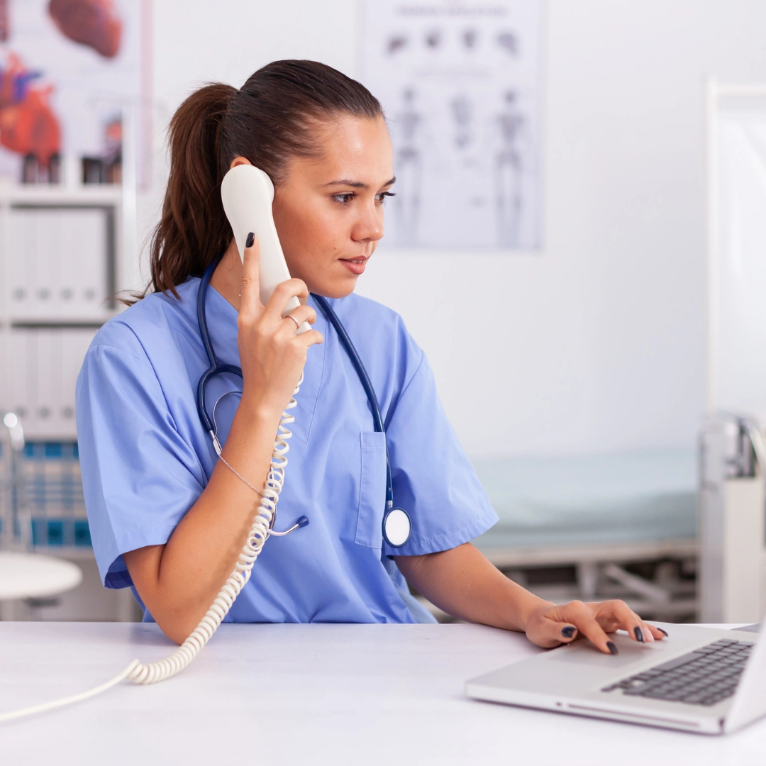 A nurse in scrubs uses a landline phone and works on a laptop at a medical office desk, assisted by a medical virtual receptionist. A nurse in scrubs uses a landline phone and works on a laptop at a medical office desk, assisted by a medical virtual receptionist.