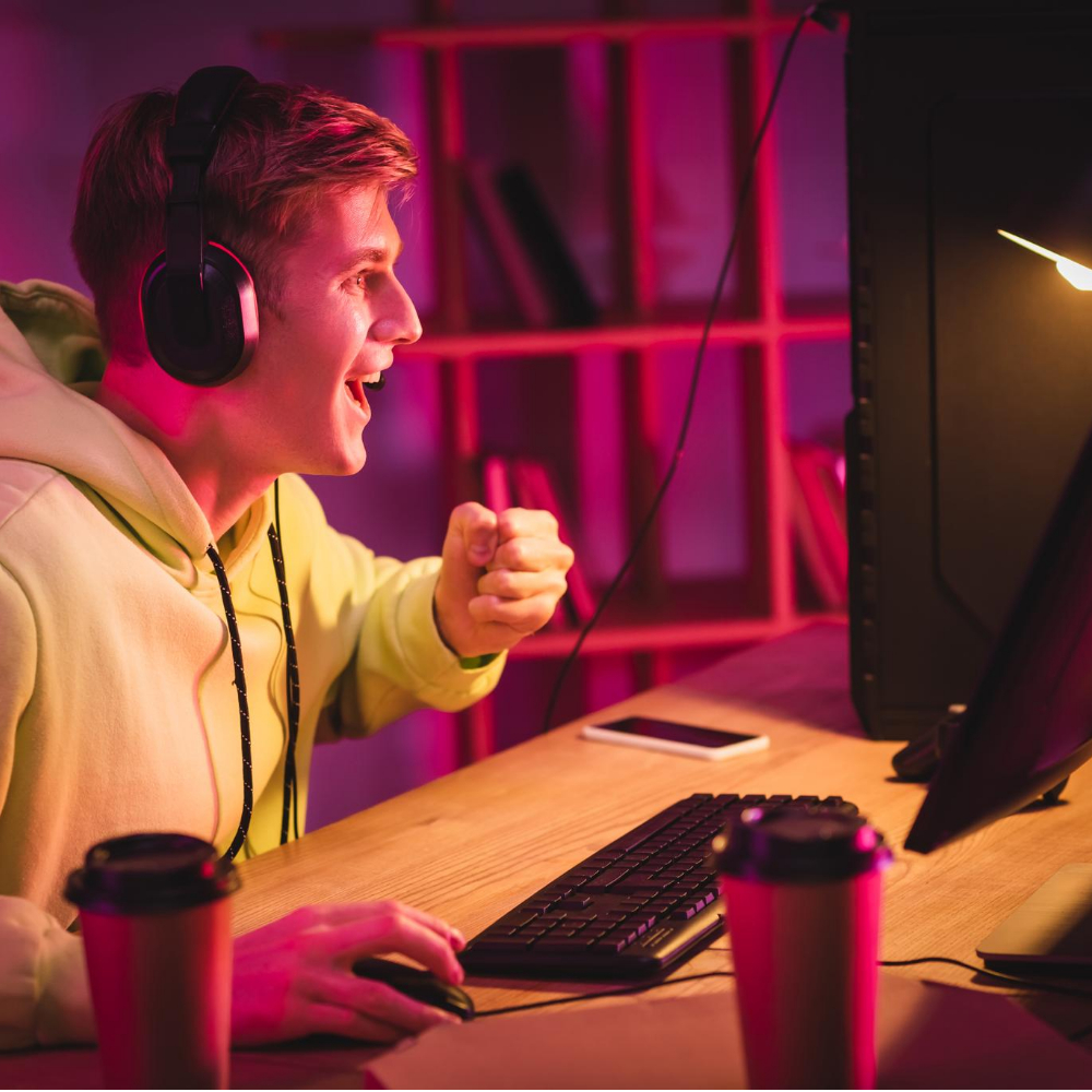 A young person wearing headphones and a hoodie sits at a desk, looking excitedly at a computer screen, surrounded by coffee cups, a keyboard, and a phone—ready to dive into gaming customer service or video game customer support tasks.