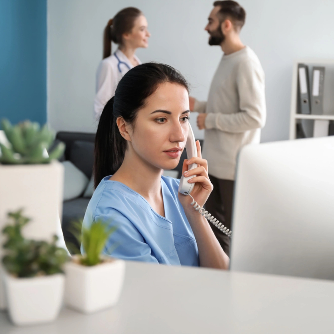 A woman in medical scrubs talks on the phone at a desk with a computer, efficiently handling healthcare appointment setting, while two people converse in the background of a modern office space.