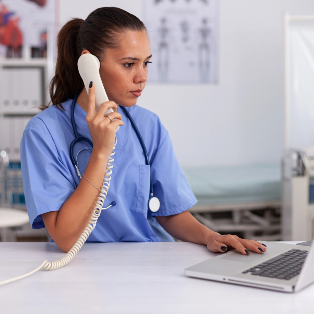 Healthcare professional in scrubs sits at a desk, talking on a landline phone and using a laptop in a medical office, possibly discussing medical billing outsourcing services.