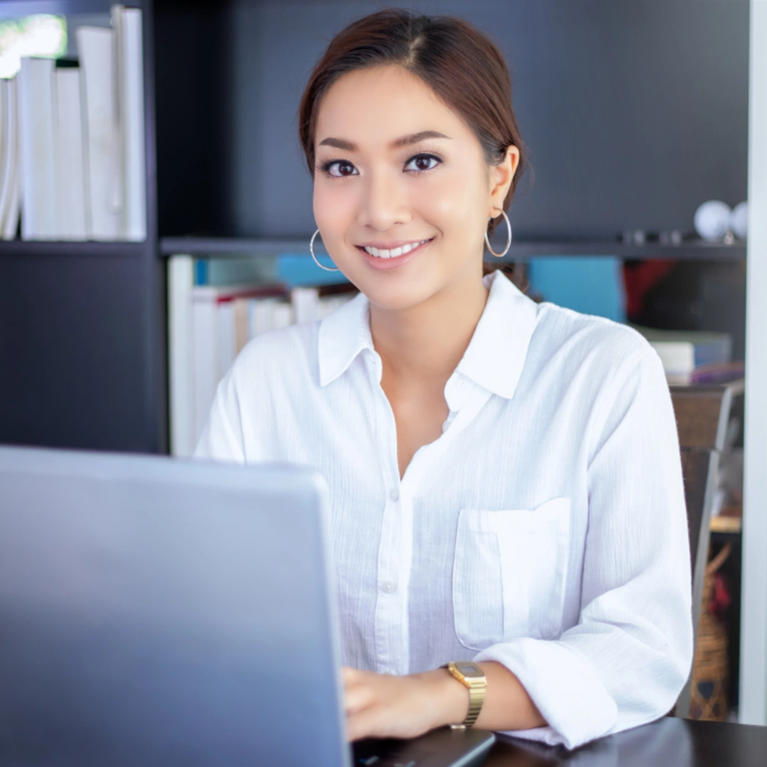 A woman in a white shirt sits at a desk, smiling while working on a laptop, illustrating the ease of managing in-app customer support with shelves of books in the background.